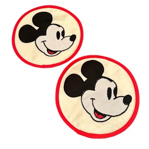 May include: Two white fabric circles with red trim, each featuring an embroidered image of Mickey Mouse's face. Mickey Mouse is smiling with black ears and eyes.