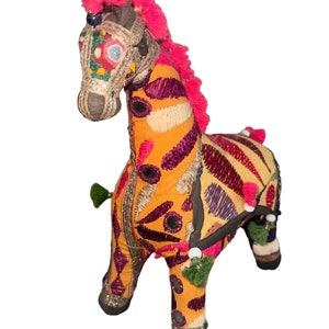May include: A decorative stuffed zebra figurine with vibrant colors. The zebra features orange, purple, and cream-colored stripes, embellished with embroidery and small mirrors. The mane and head are adorned with pink fringe and metallic accents.