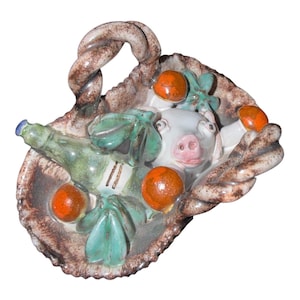 May include: A ceramic basket with a twisted handle containing a white pig figurine, a green bottle, and several orange spherical objects. The basket features a brown mottled glaze, while the decorative items include green leaves and accents.