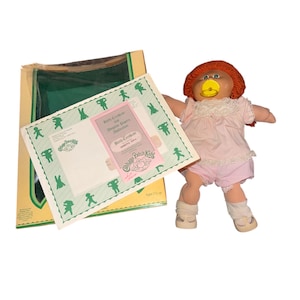 May include: A Cabbage Patch Kids doll with red yarn hair, wearing a pink dress, shorts, and white socks. The doll has a pacifier and is standing next to its original box and birth certificate.
