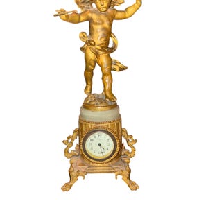 May include: A gold-colored antique clock featuring a cherub statue with outstretched arms and wings atop the clock face. The clock has a round face with Roman numerals and ornate gold detailing. The base has decorative feet.