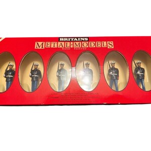 May include: A red box of Britains Metal Models, hand-painted. The box contains six miniature figurines of soldiers in blue and black uniforms, each holding a rifle. The box has a rainbow stripe on the side.