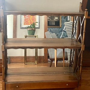 May include: A wooden shelf unit with a mirrored back and a drawer. The shelf has three shelves and a decorative X-shaped design on the sides. The shelf is brown and has a distressed finish.
