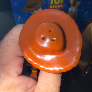 May include: Close-up of a Woody toy hat from the Disney Pixar Toy Story franchise. The hat is a vibrant orange-brown color with a rounded crown and a wide brim. The hat is held by a hand, with the Toy Story box in the background.