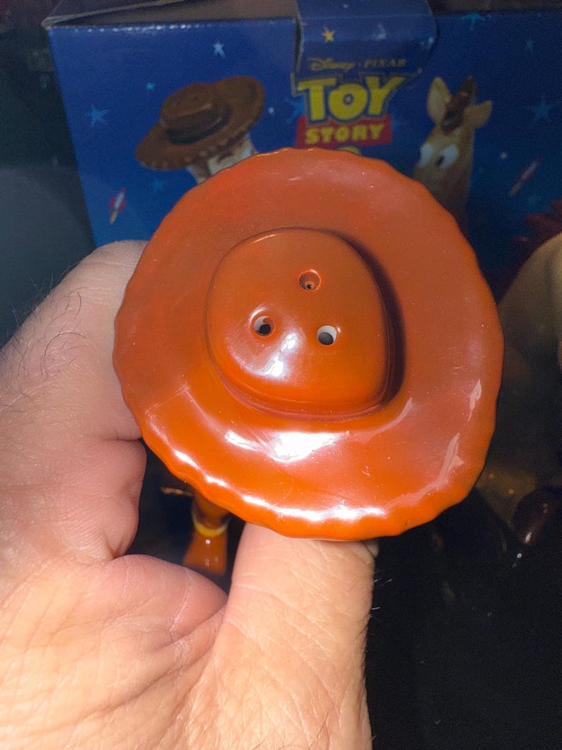 May include: Close-up of a Woody toy hat from the Disney Pixar Toy Story franchise. The hat is a vibrant orange-brown color with a rounded crown and a wide brim. The hat is held by a hand, with the Toy Story box in the background.
