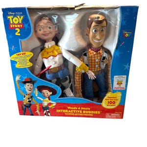 Puede incluir: Un juego de figuras de acci&oacute;n parlantes interactivas de Woody y Jessie de Toy Story 2 de Disney Pixar. La caja presenta im&aacute;genes de los personajes y el texto "Woody & Jessie INTERACTIVE BUDDIES". La caja es azul y blanca.