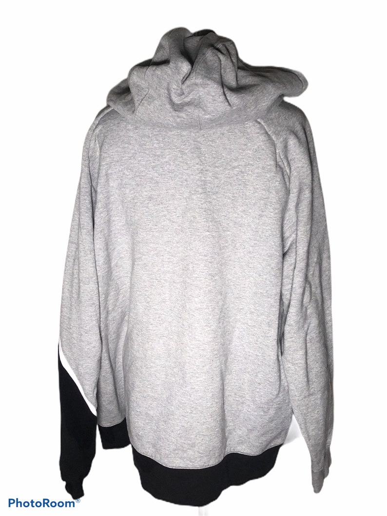 Pode incluir: Uma camisa de moletom cinza com capuz com detalhes em preto e branco. A camisa de moletom tem uma faixa preta na parte inferior e mangas pretas com detalhes em branco.