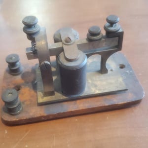 May include: Antique brass and wood telegraph key with a black metal spool. The key has multiple knobs and a spring mechanism.