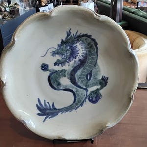 May include: A white ceramic plate with a blue and green dragon design. The plate has a scalloped edge and is on a wooden surface.