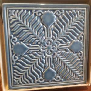 May include: A blue ceramic tile with a raised design of stylized leaves and flowers. The tile has a white border and is in a white box.