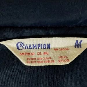 May include: A white tag with black text that reads "CHAMPION KNITWEAR CO., INC. RN 26094 100% NYLON DO NOT DRY CLEAN - DO NOT IRON EMBLEM" and a black "M" in the top right corner.