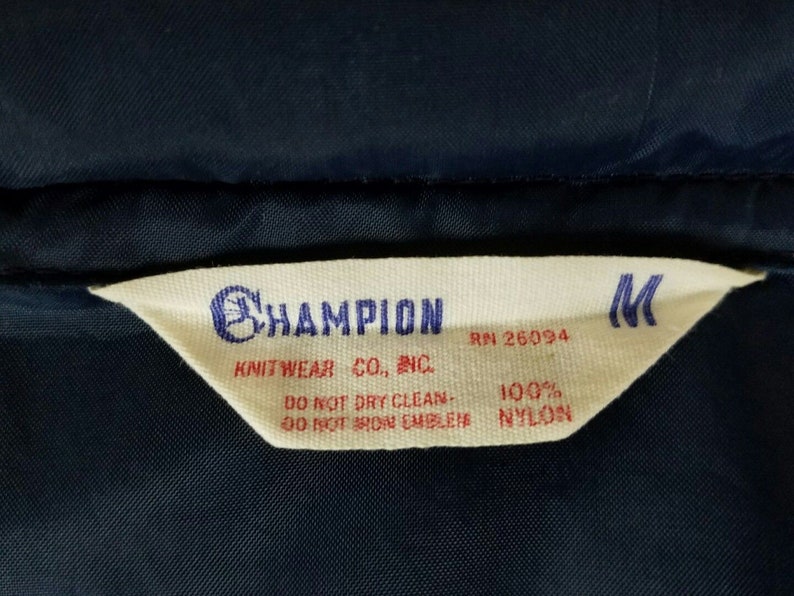 May include: A white tag with black text that reads "CHAMPION KNITWEAR CO., INC. RN 26094 100% NYLON DO NOT DRY CLEAN - DO NOT IRON EMBLEM" and a black "M" in the top right corner.