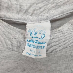 May include: A white tag with blue text that reads "Little Brownie Originals" and "Adult M" with a cartoon illustration of a smiling child. The tag is sewn onto a gray t-shirt.