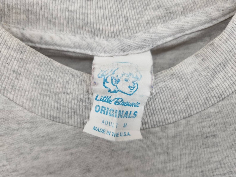 May include: A white tag with blue text that reads "Little Brownie Originals" and "Adult M" with a cartoon illustration of a smiling child. The tag is sewn onto a gray t-shirt.