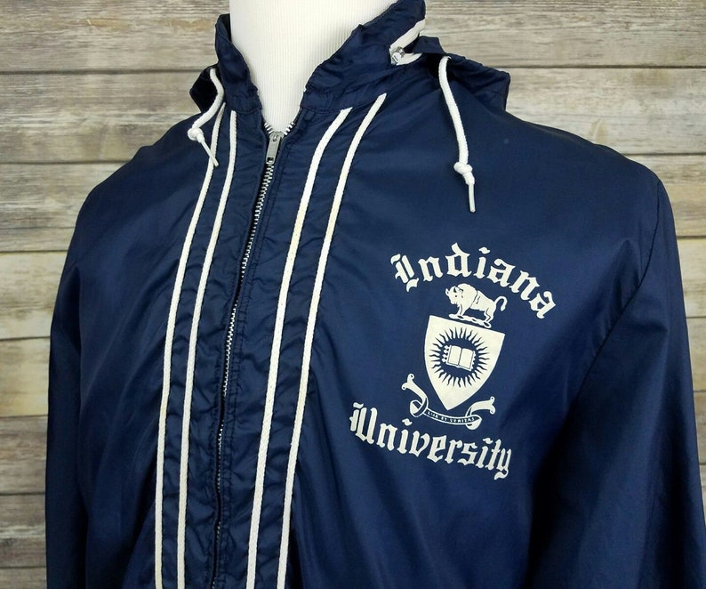 May include: A navy blue hooded jacket with white piping and a white embroidered Indiana University logo on the front. The logo features a shield with a book, a sun, and a bull.