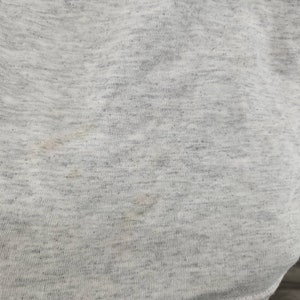 May include: Close-up of a light gray fabric with a subtle texture. The fabric has a few faint brown stains.
