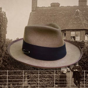 May include: A taupe felt fedora hat featuring a wide, dark navy blue grosgrain ribbon band around the crown. A small, silver-toned metal safety pin is attached to the side of the ribbon band, adding a subtle metallic detail to the hat's design.