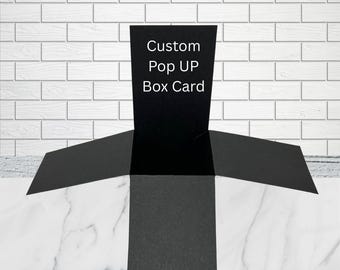 Może przedstawiać: Czarna kartka typu pop-up box o otwartej konstrukcji z czterema rozłożonymi klapkami. Na środkowym pionowym panelu znajduje się biały napis Custom Pop UP Box Card. Konstrukcja wykonana jest z matowego czarnego kartonu o geometrycznym kształcie.