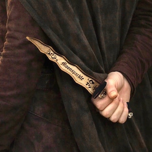 May include: A wooden dagger with the word "Moonsparkle" engraved on the blade. The dagger has a black handle and is held behind a dark brown cloak. The dagger is a fantasy-themed item.