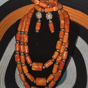 May include: A multi-strand necklace, a bracelet, and a pair of drop earrings featuring cylindrical orange marbled beads interspersed with small metallic gold-tone spherical beads. The earrings include a floral-shaped gold-tone metal stud component.