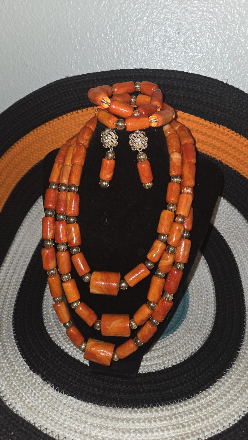 May include: A multi-strand necklace, a bracelet, and a pair of drop earrings featuring cylindrical orange marbled beads interspersed with small metallic gold-tone spherical beads. The earrings include a floral-shaped gold-tone metal stud component.