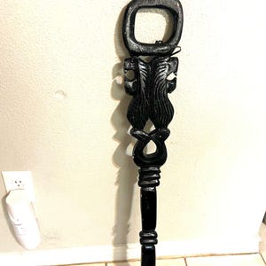 May include: A tall, black, hand-carved wooden staff featuring an open rectangular handle at the top, followed by a central section with intricate, symmetrical, textured carvings, and a smooth, tapered cylindrical shaft extending to the base.