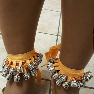 Op de afbeelding: Een paar oranje stoffen enkelbelletjes met zilveren belletjes. De belletjes zijn bevestigd aan een stoffen band die om de enkel wordt gebonden. De belletjes worden gedragen over een paar gouden sandalen.