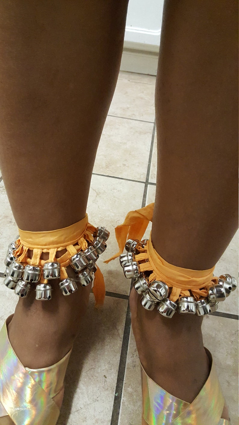 Op de afbeelding: Een paar oranje stoffen enkelbelletjes met zilveren belletjes. De belletjes zijn bevestigd aan een stoffen band die om de enkel wordt gebonden. De belletjes worden gedragen over een paar gouden sandalen.