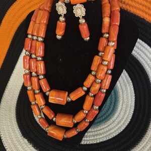 May include: A jewelry set consisting of a three-strand necklace, a bracelet, and a pair of drop earrings. The pieces feature cylindrical orange beads interspersed with small gold-toned spherical beads. The earrings have gold-toned flower studs.