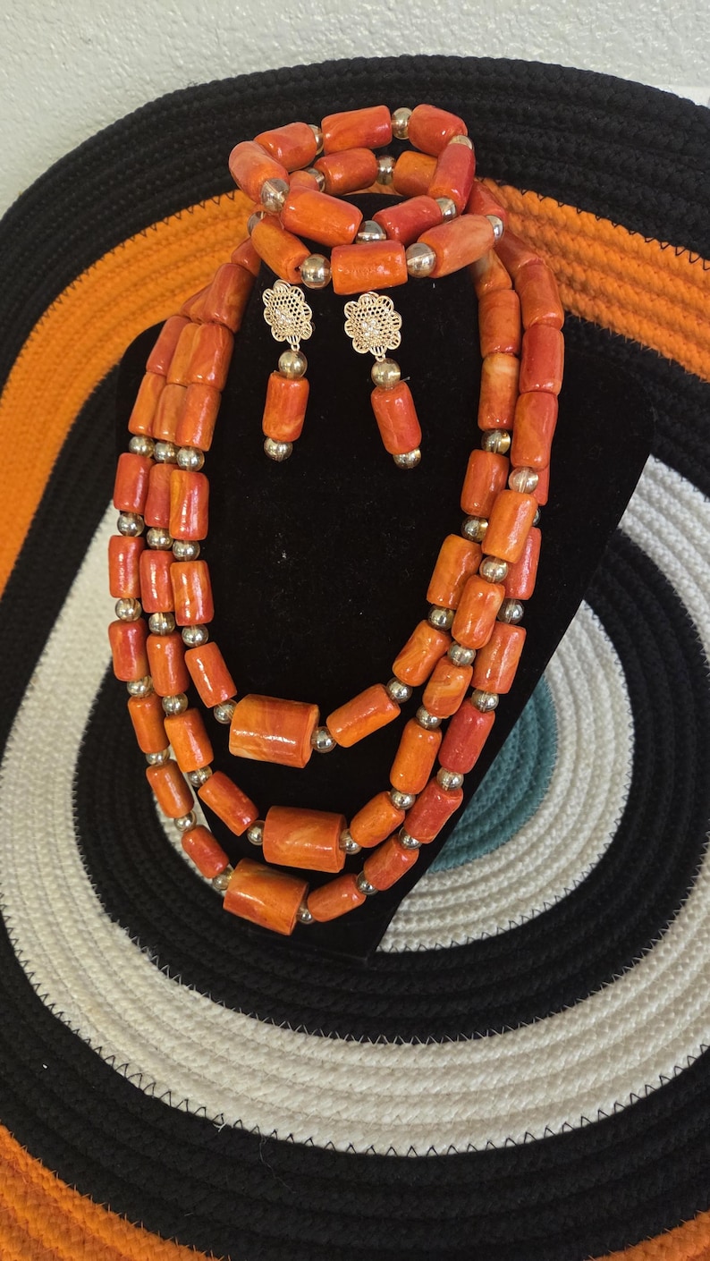 May include: A jewelry set consisting of a three-strand necklace, a bracelet, and a pair of drop earrings. The pieces feature cylindrical orange beads interspersed with small gold-toned spherical beads. The earrings have gold-toned flower studs.