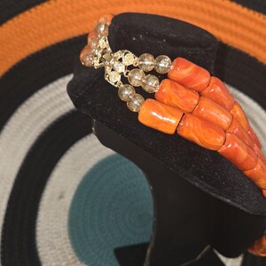 May include: A multi-strand necklace featuring three rows of cylindrical orange beads with marbled patterns, interspersed with clear, round faceted beads. The strands are connected by a gold-toned metal clasp with intricate filigree detailing.