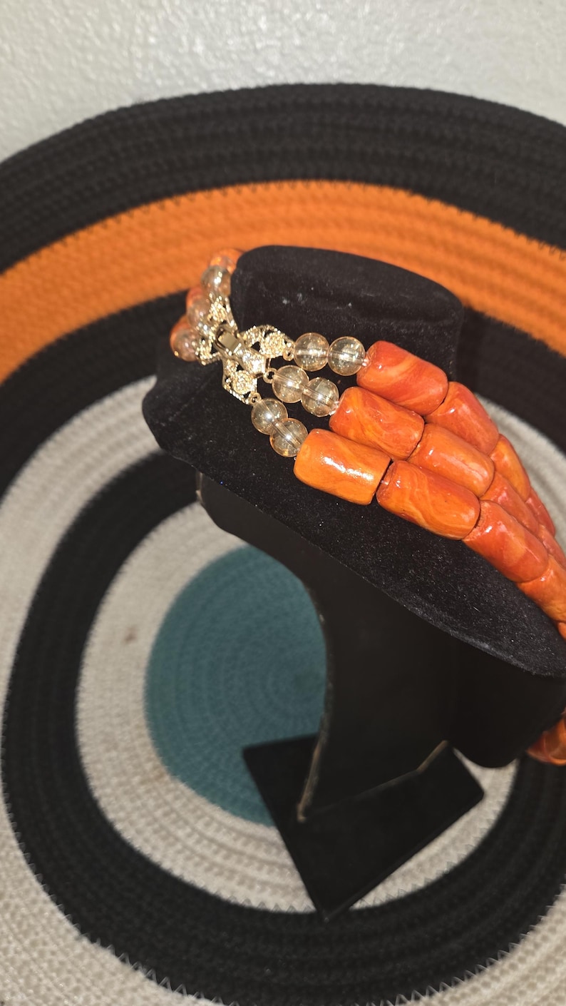 May include: A multi-strand necklace featuring three rows of cylindrical orange beads with marbled patterns, interspersed with clear, round faceted beads. The strands are connected by a gold-toned metal clasp with intricate filigree detailing.
