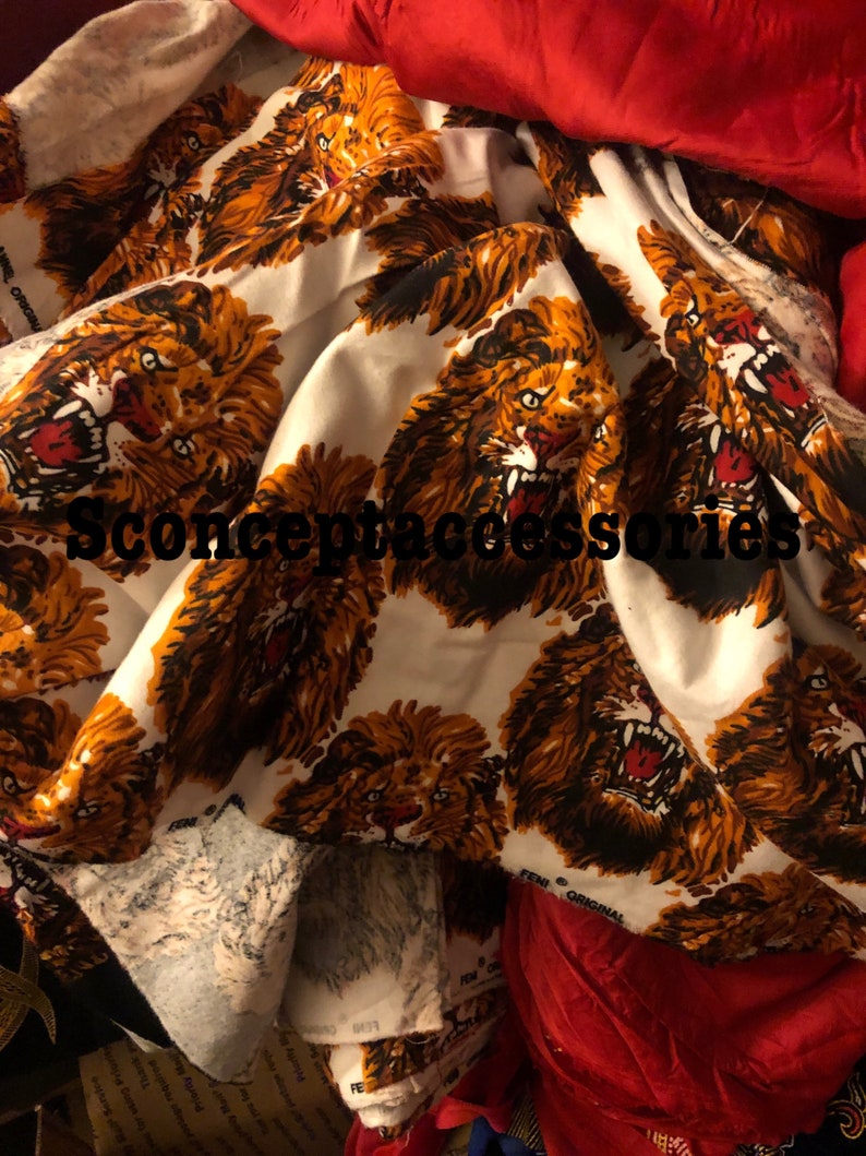May include: White fabric with a repeating pattern of orange and brown lions with black outlines. The lions have open mouths and sharp teeth. The fabric is folded and crumpled.