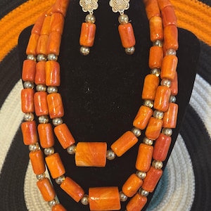 May include: A multi-strand necklace, a bracelet, and a pair of drop earrings featuring cylindrical orange beads with marbled patterns, interspersed with small metallic gold-tone spherical beads. The earrings have gold-tone flower-shaped studs.