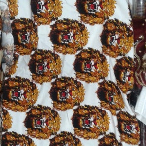 May include: White fabric with a repeating pattern of brown and black lion faces with red mouths. The lion faces are printed on a white background.