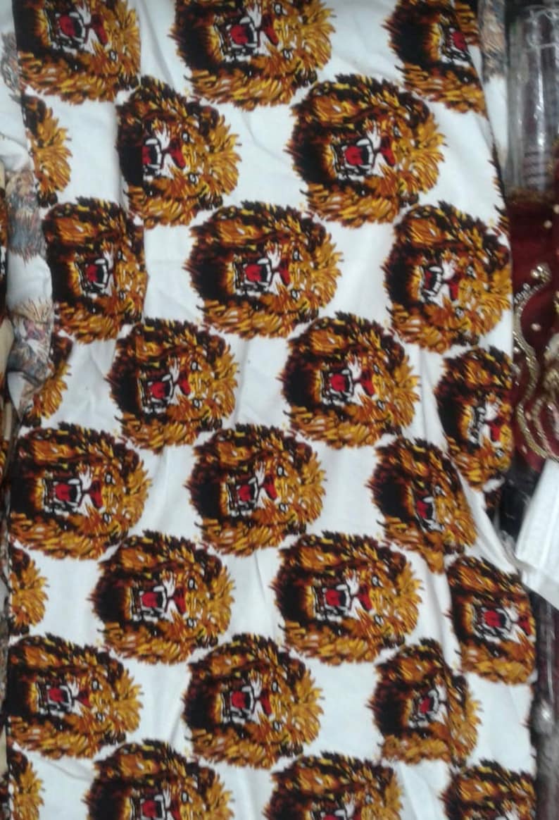 May include: White fabric with a repeating pattern of brown and black lion faces with red mouths. The lion faces are printed on a white background.