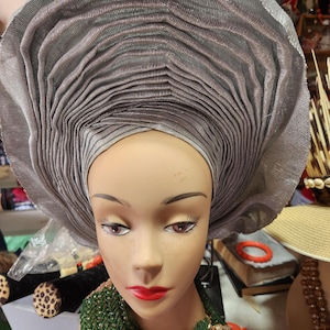Può includere: Un gele grigio argento, un tradizionale copricapo nigeriano, è esposto su una testa di manichino. Il gele è finemente pieghettato e stratificato, creando una forma voluminosa a ventaglio. Il manichino ha rossetto rosso e una collana di perline verdi.