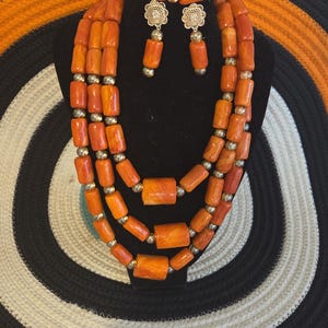 May include: A multi-strand necklace, a bracelet, and a pair of earrings made of orange cylindrical beads interspersed with small gold-toned spherical beads. The earrings feature a gold-toned floral filigree stud with a single dangling orange bead.
