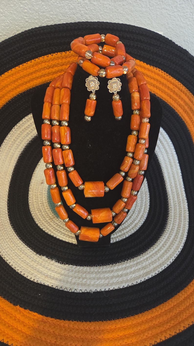 May include: A multi-strand necklace, a bracelet, and a pair of earrings made of orange cylindrical beads interspersed with small gold-toned spherical beads. The earrings feature a gold-toned floral filigree stud with a single dangling orange bead.