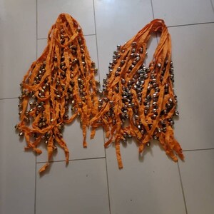 Op de afbeelding: Twee oranje stoffen stroken met rijen kleine zilveren belletjes. De stroken zijn aan de bovenkant aan elkaar gebonden.