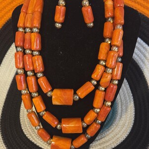 May include: A three-strand necklace and matching bracelet set featuring cylindrical orange marbled beads interspersed with small metallic gold spheres. The set includes a pair of drop earrings with gold-tone floral filigree studs.