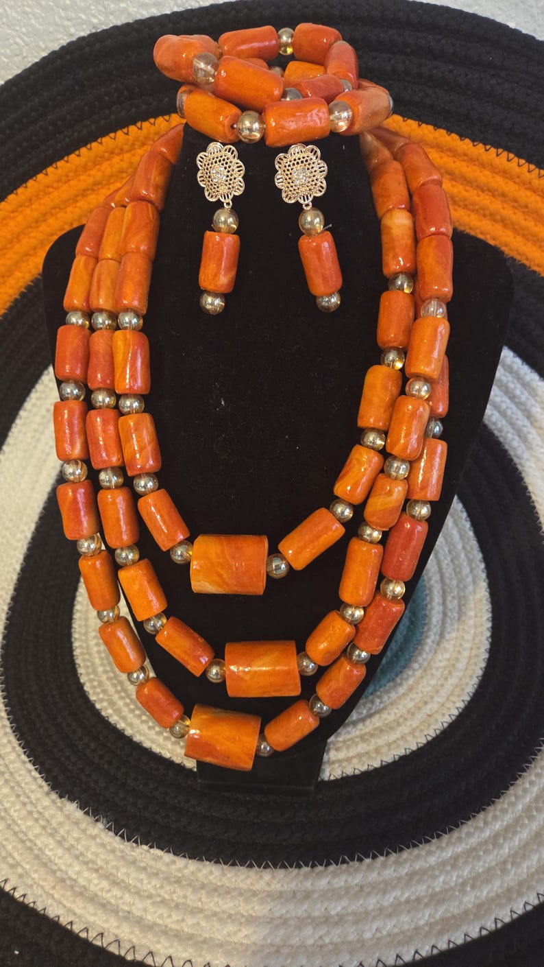 May include: A three-strand necklace and matching bracelet set featuring cylindrical orange marbled beads interspersed with small metallic gold spheres. The set includes a pair of drop earrings with gold-tone floral filigree studs.