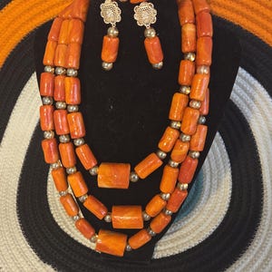 May include: A multi-strand necklace, a bracelet, and a pair of drop earrings featuring cylindrical orange beads with marbled patterns, interspersed with small metallic gold-toned spherical beads. The earrings include a gold-toned floral stud component.