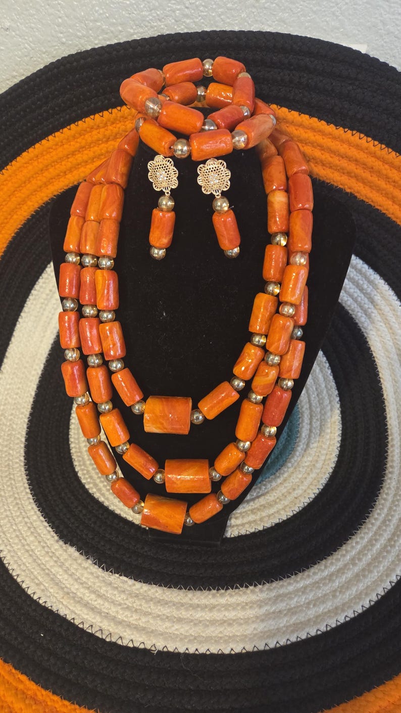 May include: A multi-strand necklace, a bracelet, and a pair of drop earrings featuring cylindrical orange beads with marbled patterns, interspersed with small metallic gold-toned spherical beads. The earrings include a gold-toned floral stud component.