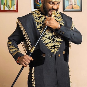 May include: Black formal suit with gold embroidery and trim, paired with a gold hat. The suit features a unique design with leaf-like gold patterns and decorative tassels. A black cane with gold accents completes the ensemble.