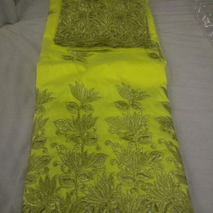 Puede incluir: Tela amarillo brillante con bordado floral dorado. La tela est&aacute; doblada, revelando una secci&oacute;n del borde bordado y un trozo m&aacute;s peque&ntilde;o de la misma tela. El bordado presenta patrones florales detallados, creando un aspecto lujoso.