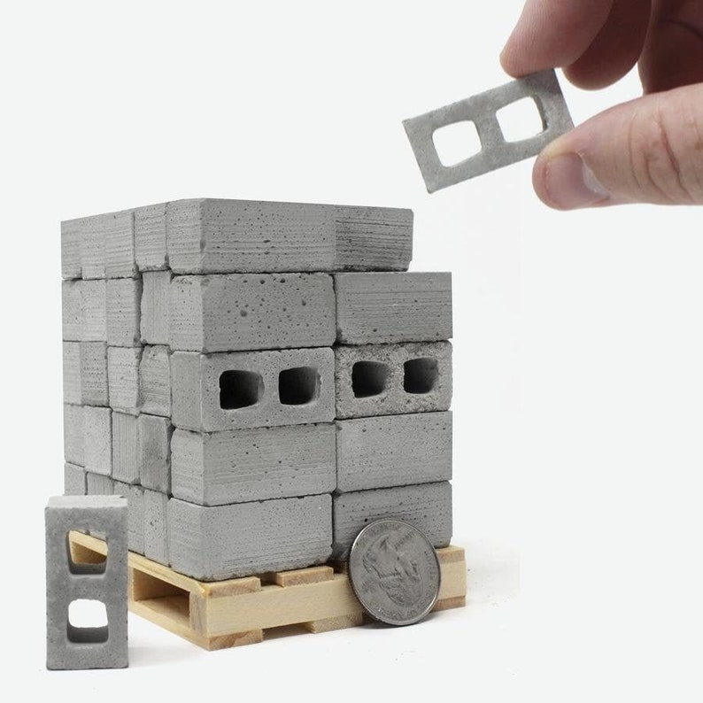 May include: A stack of miniature concrete blocks on a wooden pallet. The blocks are gray and have a rough texture. A single block is being held by a hand, and a US quarter is placed next to the pallet for scale.