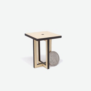May include: A small, square wooden table with a black painted edge. The table has a simple, modern design with a cross-shaped base.