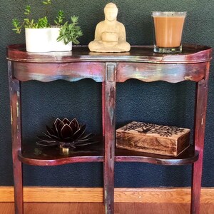 May include: A distressed wooden side table with two shelves. The top shelf has a small Buddha statue and a glass of brown liquid. The bottom shelf has a metal lotus flower and a wooden box with a tree design.