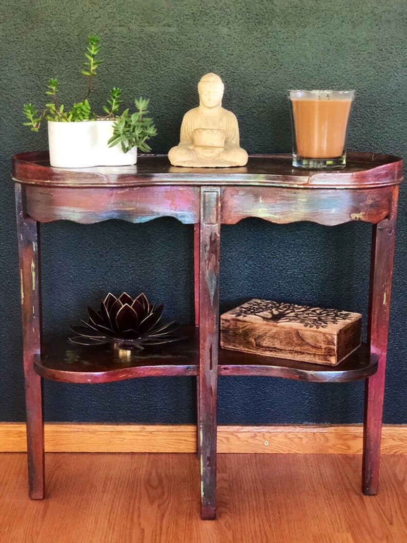 May include: A distressed wooden side table with two shelves. The top shelf has a small Buddha statue and a glass of brown liquid. The bottom shelf has a metal lotus flower and a wooden box with a tree design.